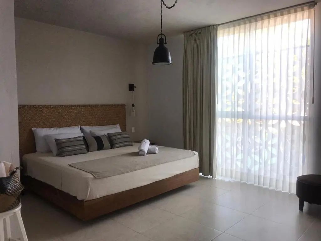 Hell gestaltetes Schlafzimmer mit Doppelbett, geflochtenem Kopfteil und Fensterfront.