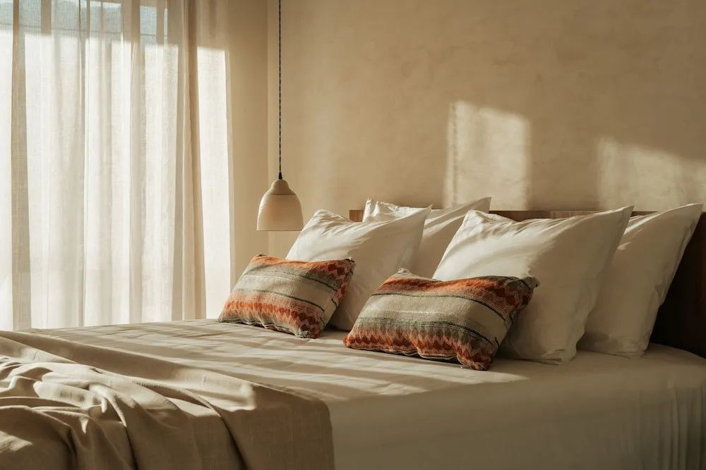 Doppelbett mit weißen Kissen, gemusterten Decken und einer hängenden Lampe im Sonnenlicht.