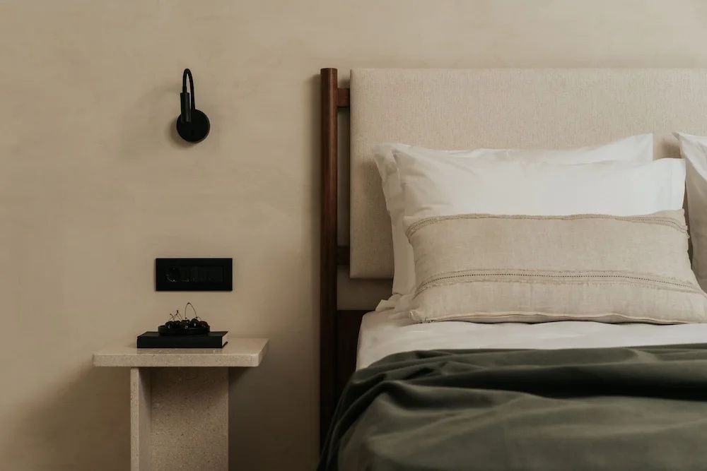Bett mit gepolstertem Kopfteil, Nachttisch, schwarzer Wandlampe und weißer Bettwäsche.
