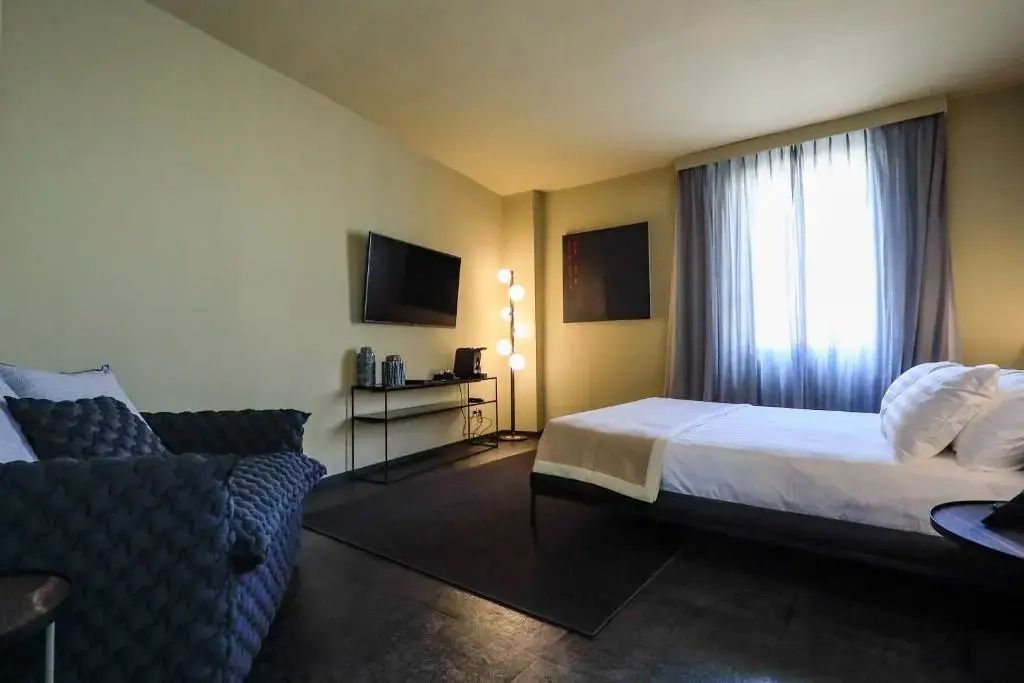 Hotelzimmer mit Wandfernseher, moderner Stehlampe und dunklem Teppich.