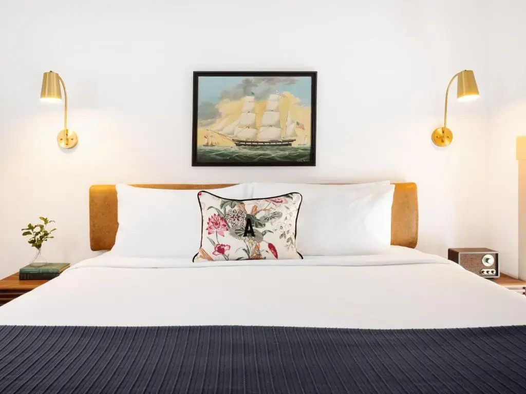 Doppelbett mit weißer Bettwäsche, buntem Kissen und Schiffsbild an der Wand.