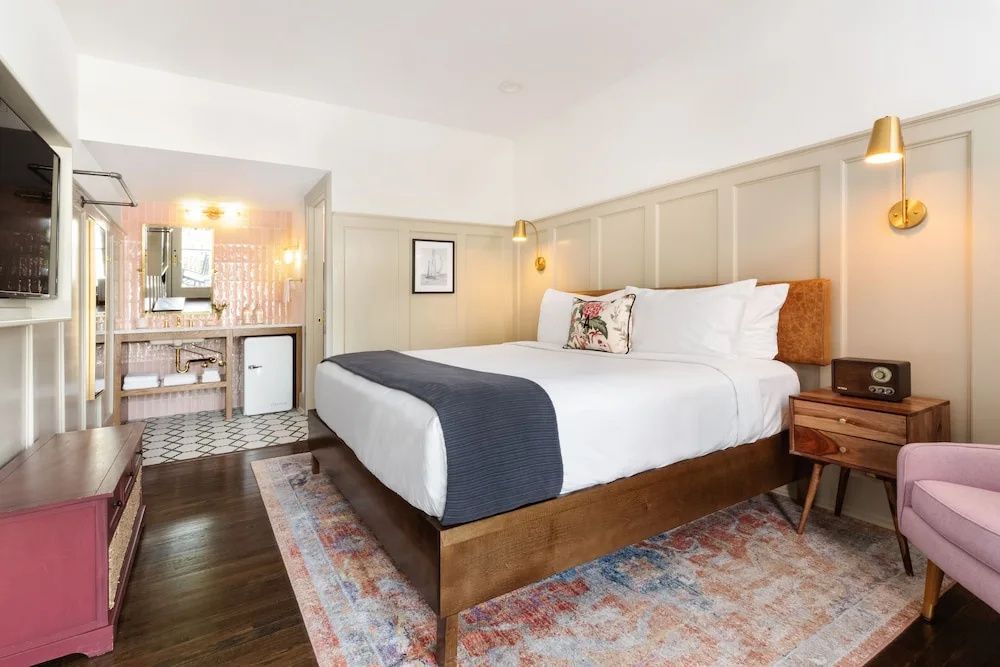 Hotelzimmer mit Parkettboden, buntem Teppich, großem Bett und Blick ins Bad.
