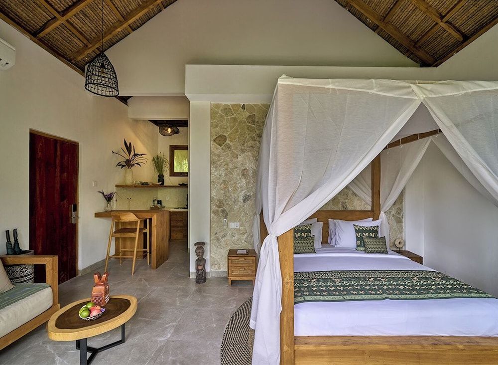 Schlafzimmer mit hohem Reetdach, Himmelbett und einer Küchenzeile aus Holz im Hintergrund.
