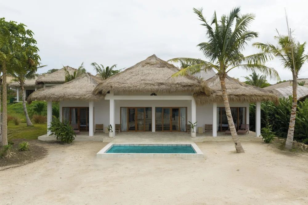 Reetgedeckte Villa mit kleinem Pool auf Sandboden und einer Palme vor der Fassade.