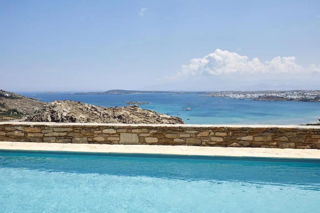 Infinitypool mit Blick auf das blaue Meer und die hügelige Küste unter blauem Himmel.
