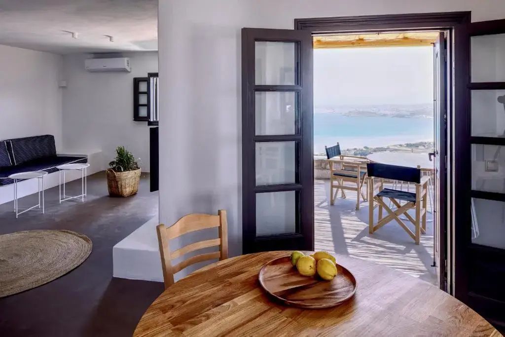 Blick durch die geöffnete Tür auf eine Terrasse mit Holzstühlen und Ausblick auf das Meer.