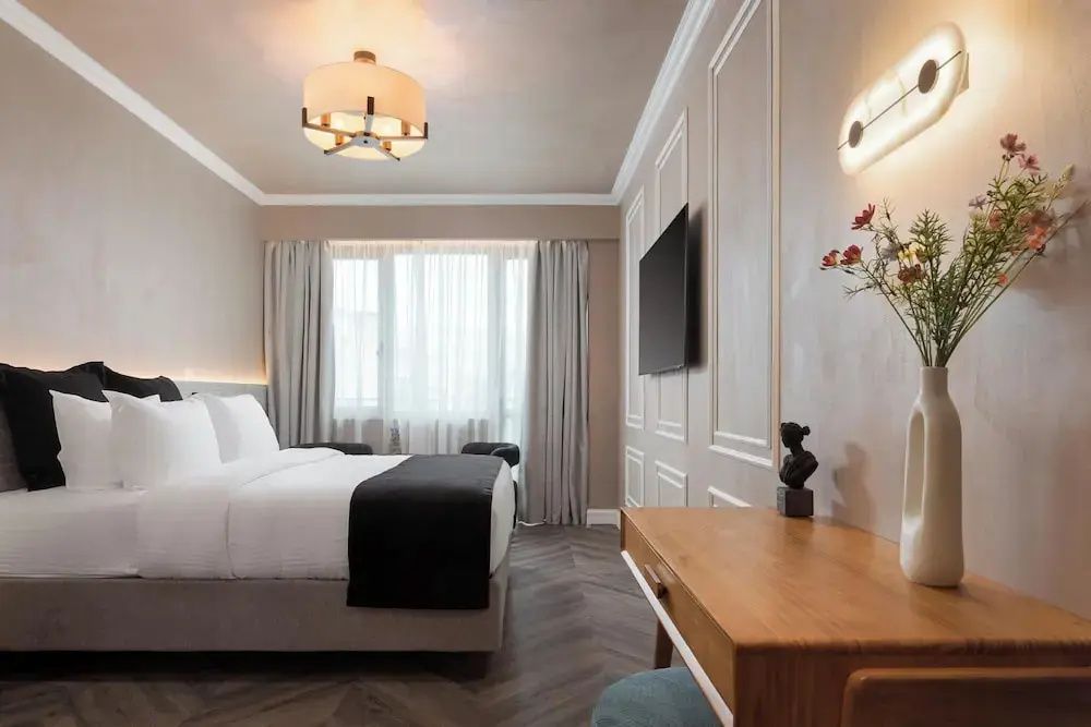 Schlafzimmer mit Doppelbett, Wandfernseher, Holzschreibtisch und hellem Fenster mit Vorhängen.