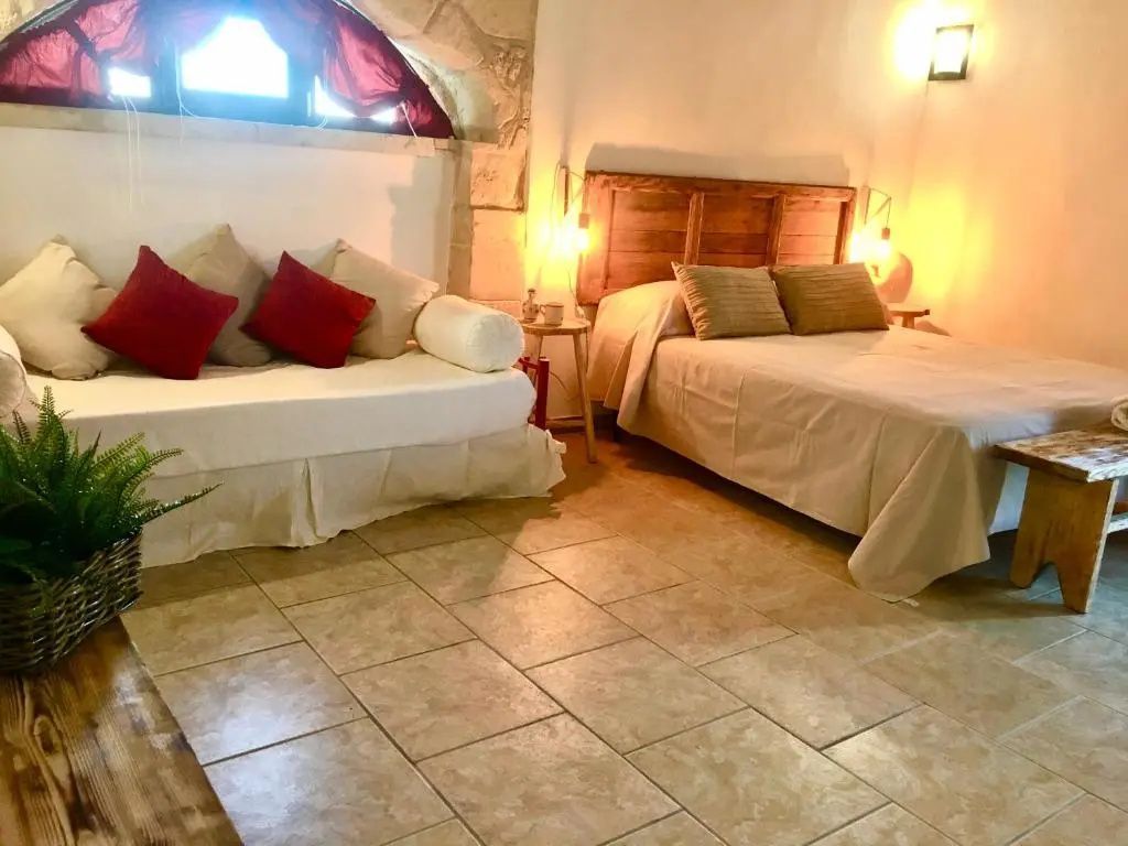 Schlafzimmer mit Doppelbett, Tagesbett und rotem Kissen unter einem kleinen Steinbogen.