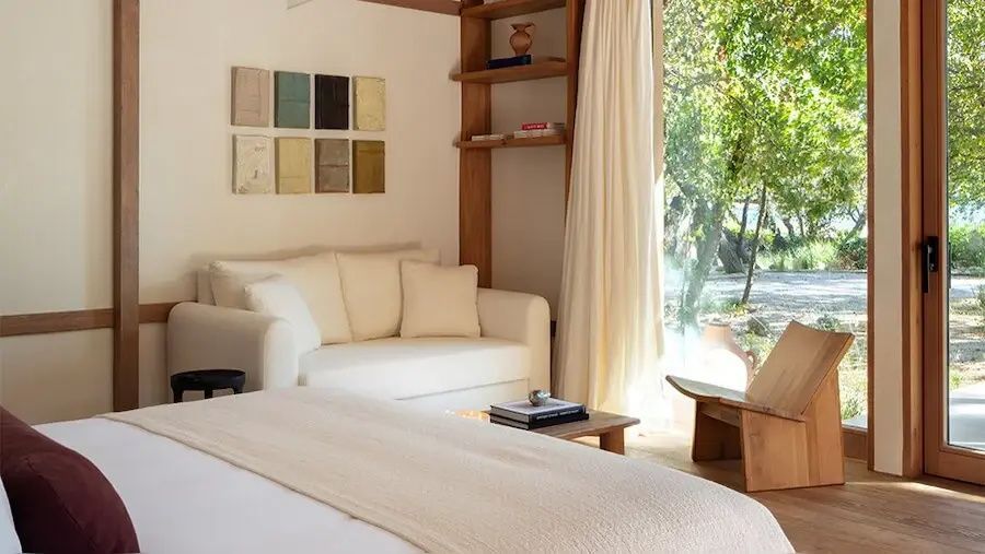 Helles Hotelzimmer mit weißem Sofa, Holzregalen und Blick durch große Fenster ins Grüne.