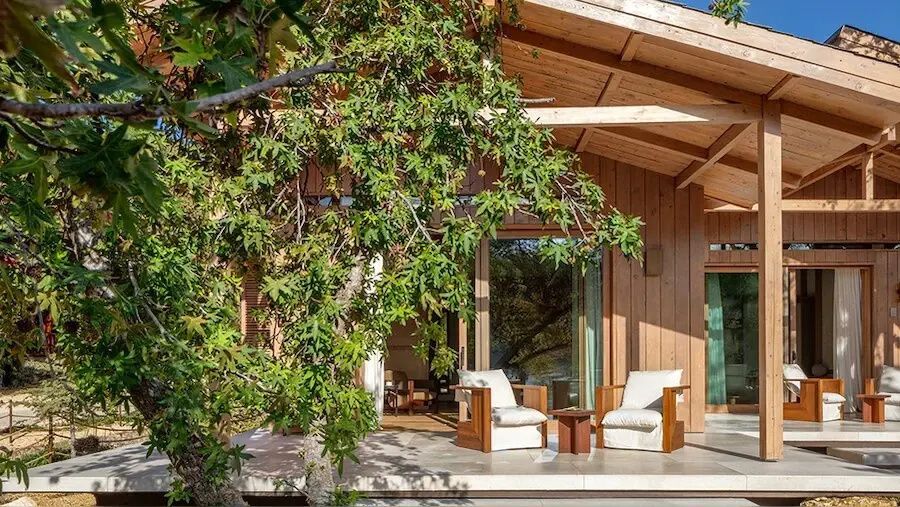 Überdachte Holzterrasse mit zwei Sesseln und einem kleinen Tisch vor Bäumen.