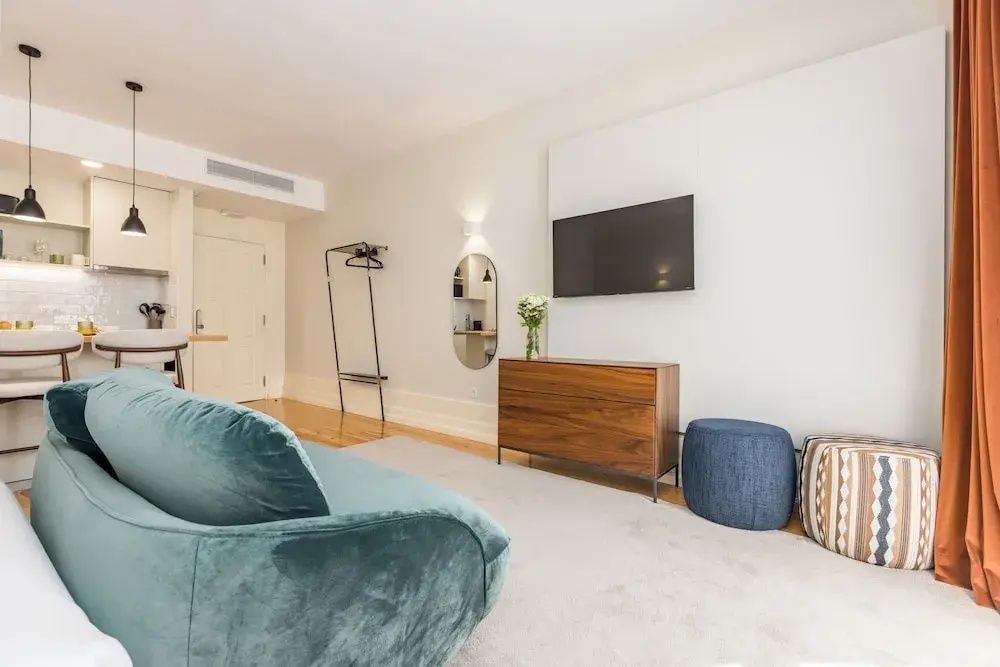 Sitzbereich im Zimmer mit Holzkommode, Fernseher, blauem Hocker und Kleiderständer.