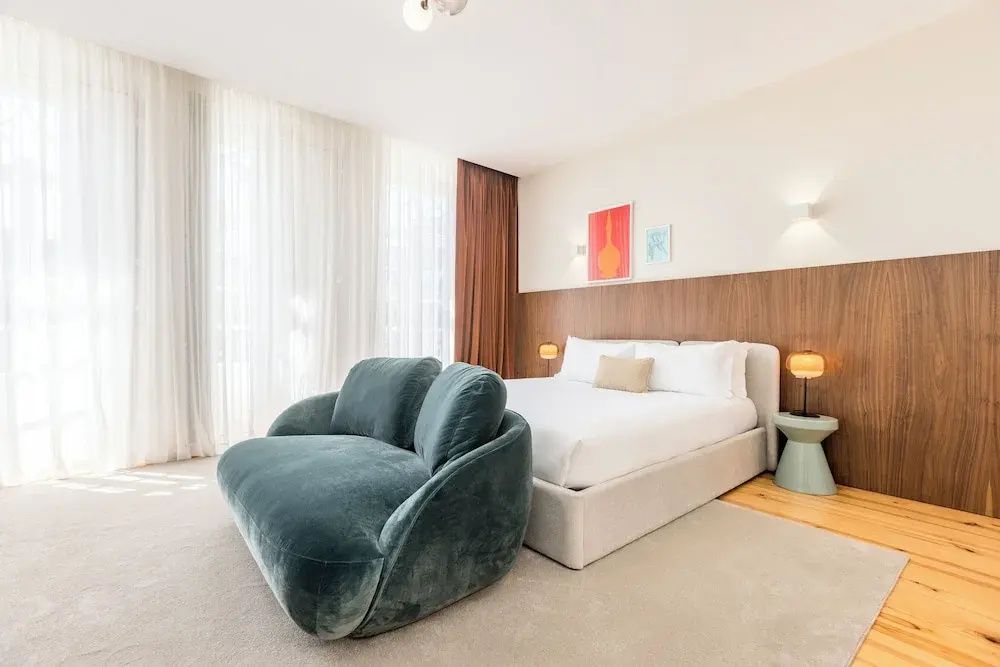 Offenes Zimmer mit großem grünem Samtsessel im Vordergrund und Doppelbett im Hintergrund.