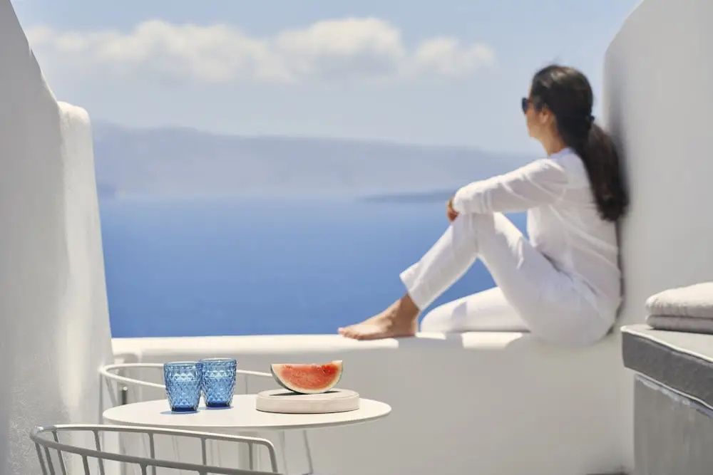 Frau in weißer Kleidung sitzt auf einer weißen Balkonmauer mit Blick auf das blaue Meer.