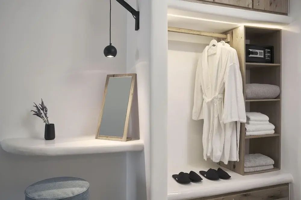Weißer Schminkplatz mit Holzspiegel und offener Kleiderschrank mit Bademantel und Hausschuhen.