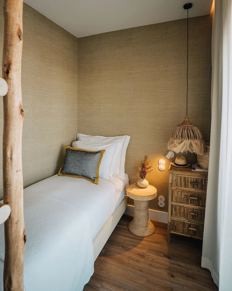 Einzelbett mit weißer Bettwäsche neben einer Holzkommode und geflochtener Lampe.