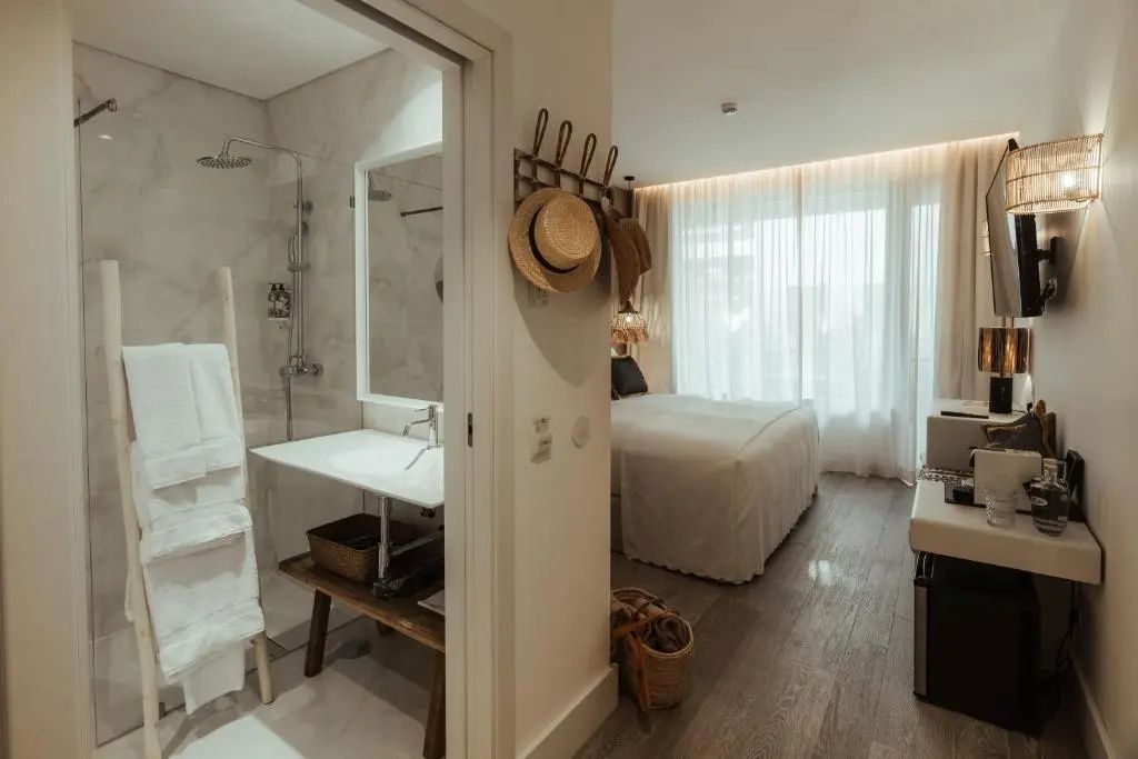 Blick in ein Hotelzimmer mit angrenzendem Badezimmer, weißem Doppelbett und großem Fenster mit hellen Vorhängen.