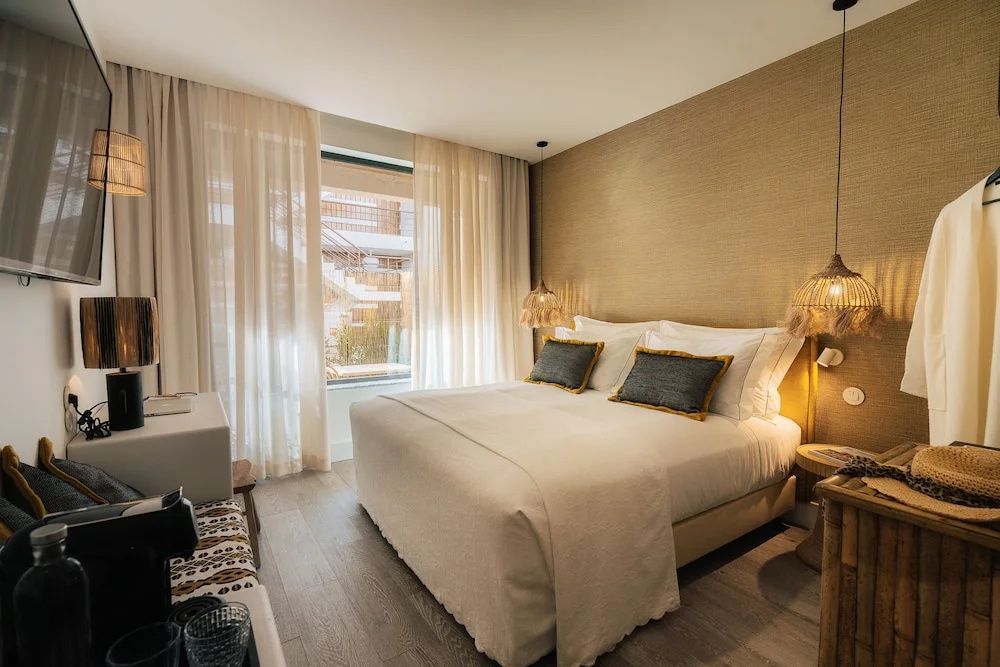 Doppelbett mit weißer Bettwäsche und grauen Kissen in einem hell eingerichteten Hotelzimmer mit Vorhängen.