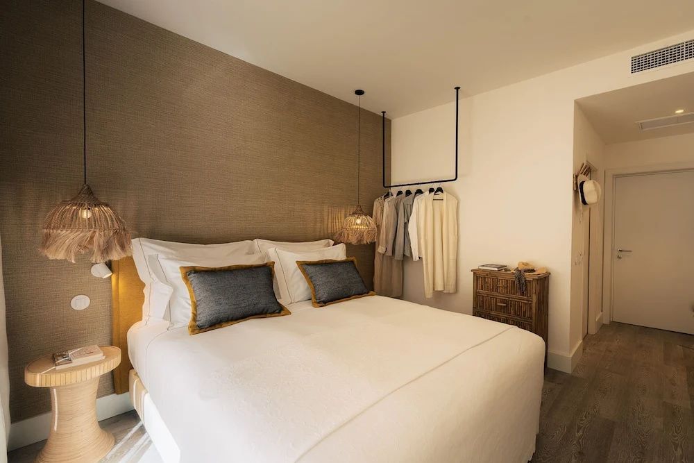 Doppelbett vor beigefarbener Wand mit Hängeleuchte und Kleiderstange im Schlafbereich eines Hotelzimmers.