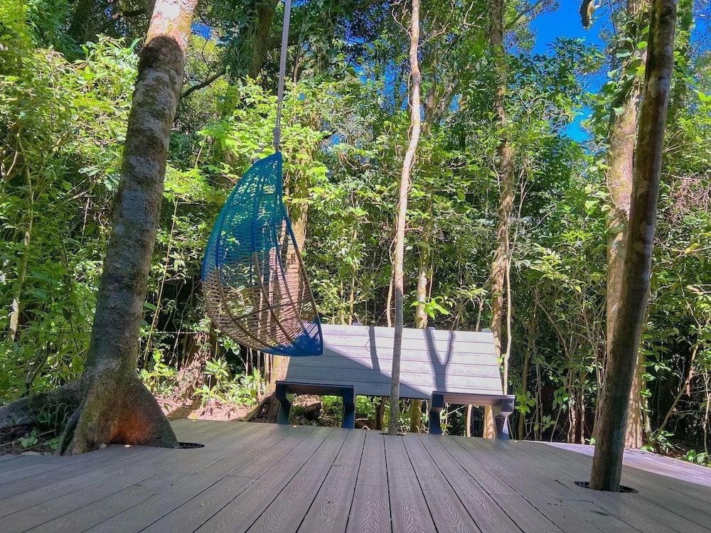 Holzdeck im Grünen mit einem blauen Hängesessel und einer Holzbank zwischen Bäumen.