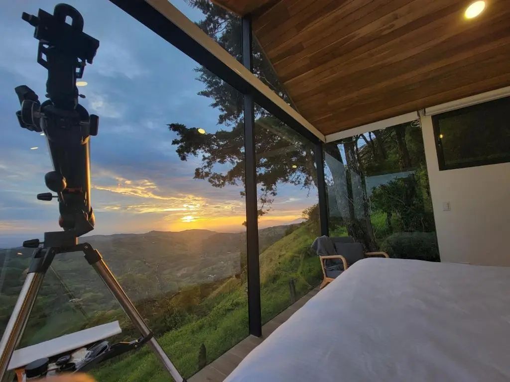 Blick durch ein großes Fenster bei Sonnenuntergang über eine hügelige Landschaft.