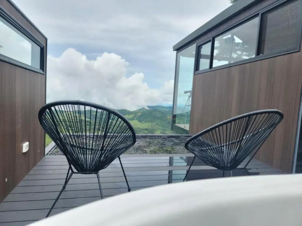 Zwei schwarze Stühle auf einer Holzterrasse mit Blick auf grüne Hügel und Wolken.