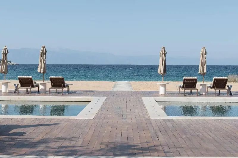 Symmetrische Poolanlage auf einer Holzterrasse mit Liegestühlen direkt am Sandstrand und Meer.
