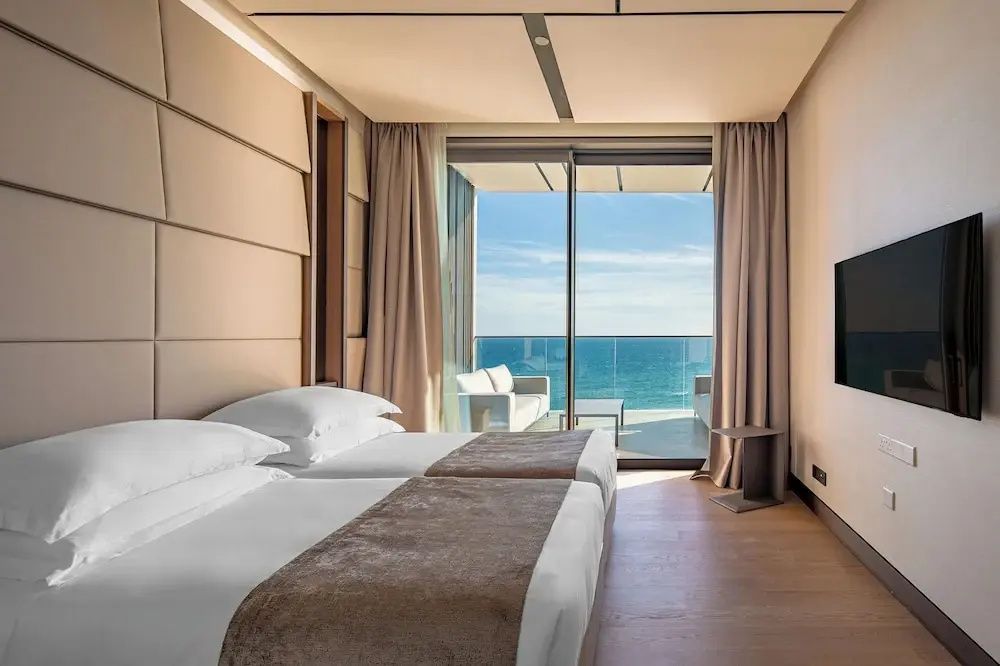 Schlafzimmer mit zwei Betten und Glasflügeltür zum Balkon mit Meerblick.