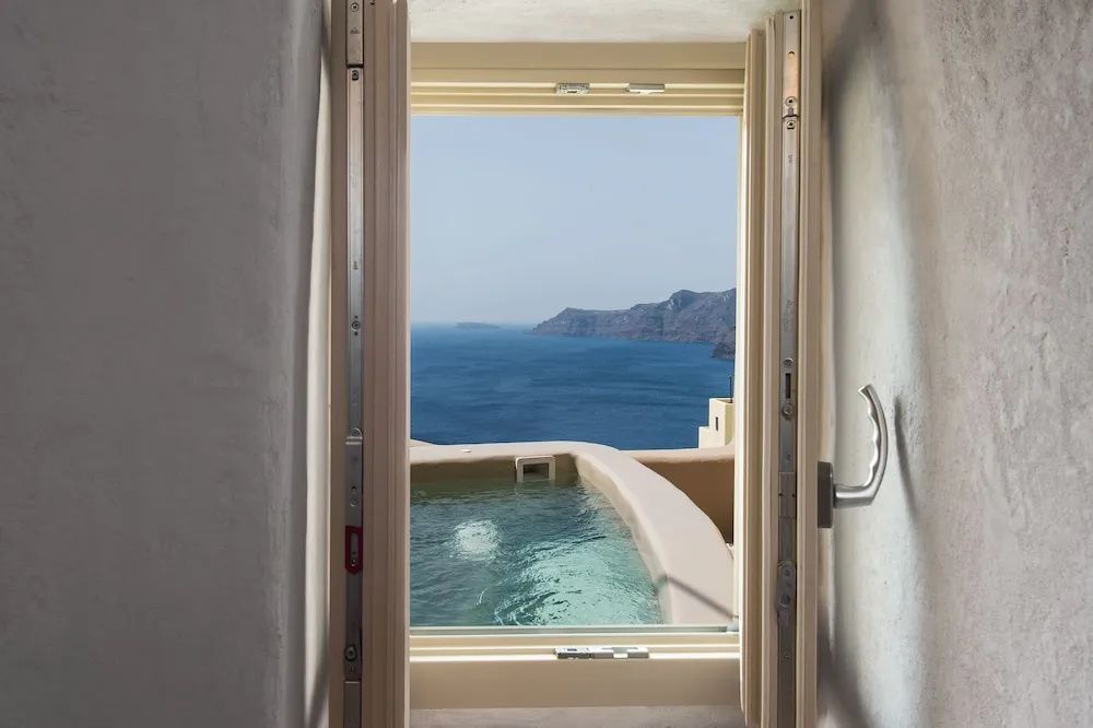 Blick durch ein geöffnetes Fenster auf ein Außenbecken und das dahinterliegende Meer.