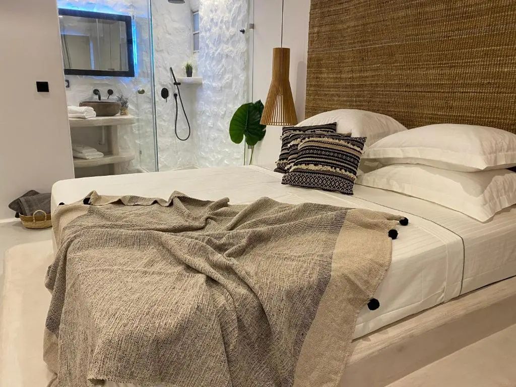 Doppelbett mit gewebtem Kopfteil, Kissen und Decke vor einer transparenten Duschkabine.