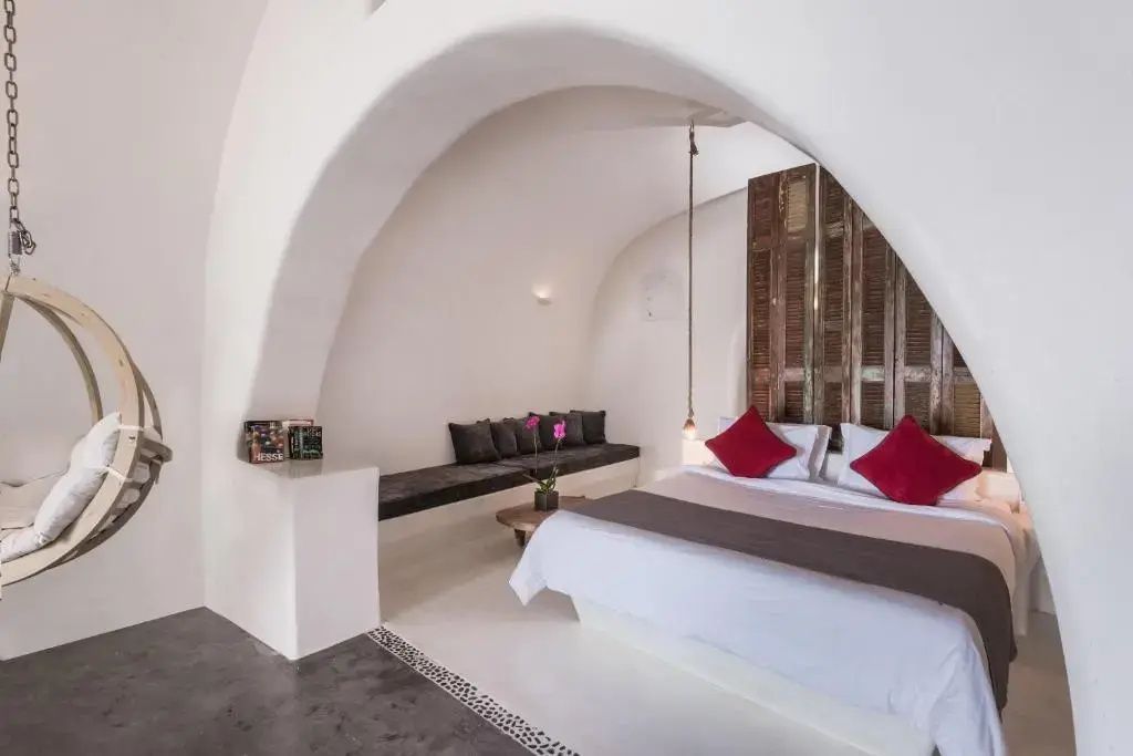 Weiß gewölbtes Hotelzimmer mit Doppelbett, roten Kissen und eingebauter Sitzbank an der Wand.
