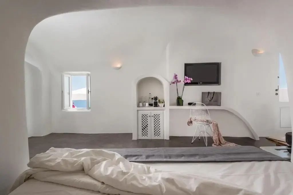 Blick über das Bett auf die weiße Zimmerwand mit Fernseher, Schreibplatz und kleinem Fenster.