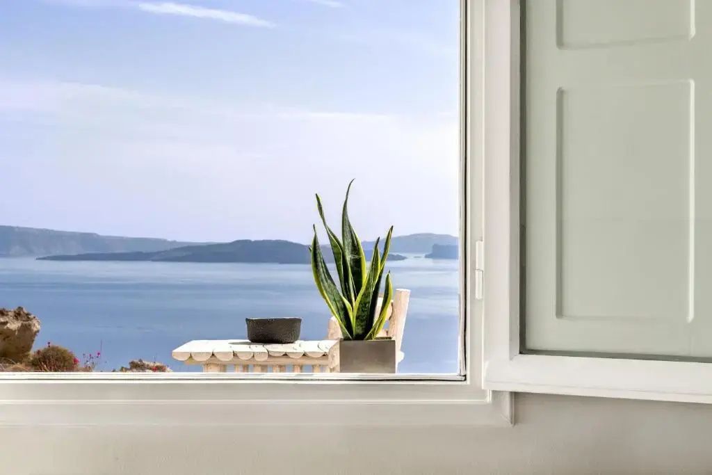 Blick durch ein offenes Fenster mit Zimmerpflanze auf das Meer und gegenüberliegende Inseln.
