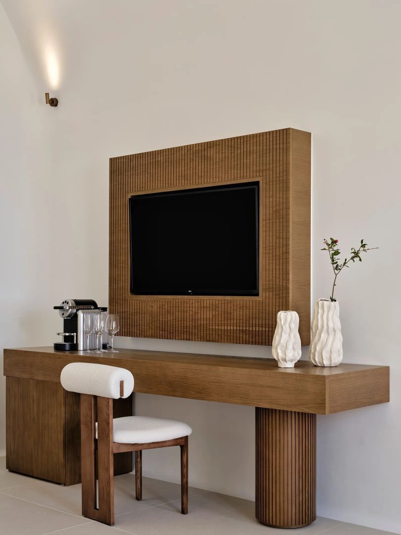 Holzschreibtisch mit Wand-Fernseher, Kaffeemaschine, Gläsern und Dekorationsvasen.