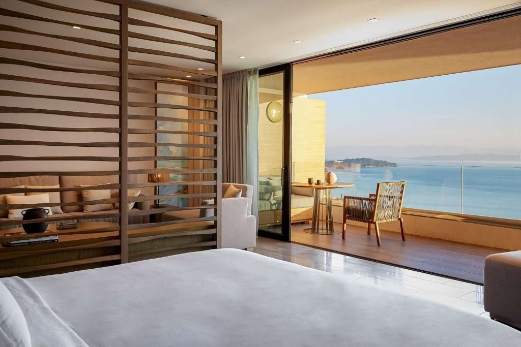 Blick vom Hotelzimmer über einen möblierten Balkon auf das blaue Meer und die Küste.