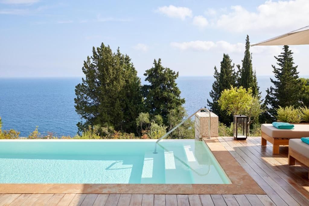 Pool mit Einstiegstreppe direkt am Hang mit freier Aussicht auf das blaue Meer und grüne Zypressen.