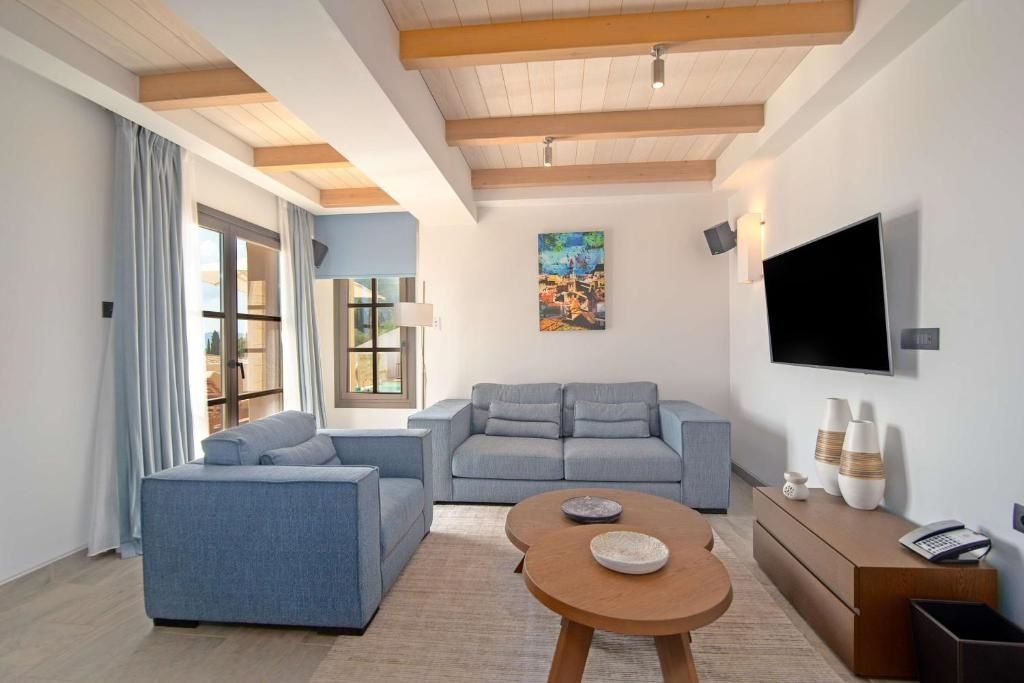 Wohnbereich einer Suite mit blauen Sofas, runden Holztischen und Fernseher an der Wand.