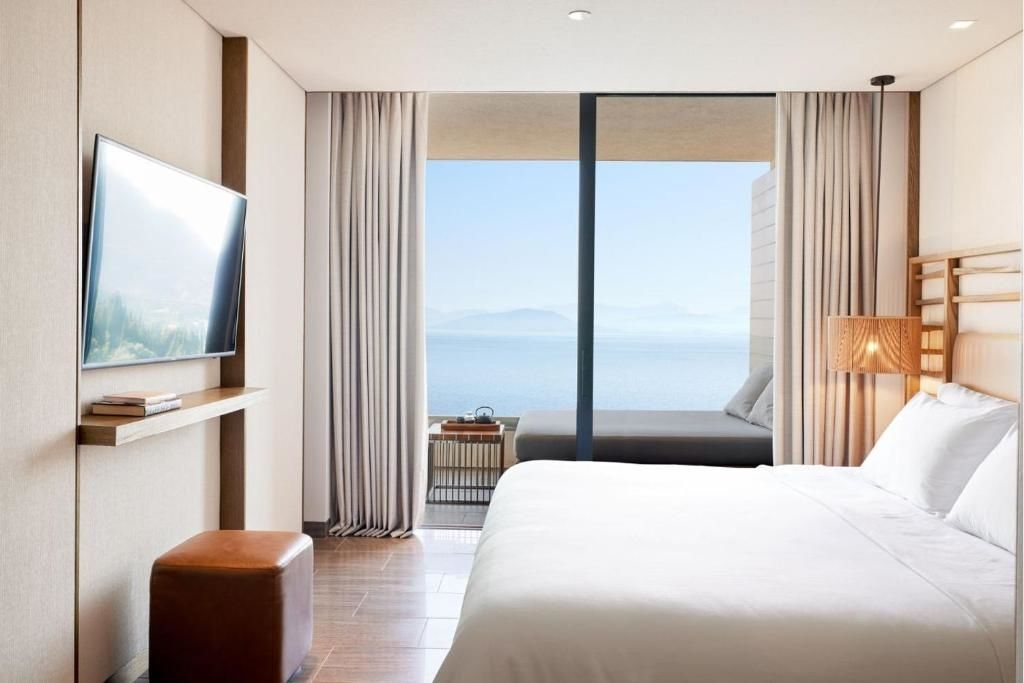 Schlafzimmer mit hellem Doppelbett und großem Fensterfront-Zugang zum Balkon mit Meeresblick.