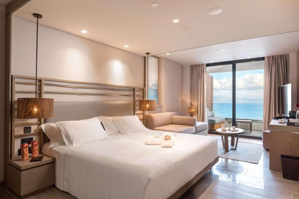Helles Hotelzimmer mit großem Bett, Couch und Glasfront mit Blick auf das Meer