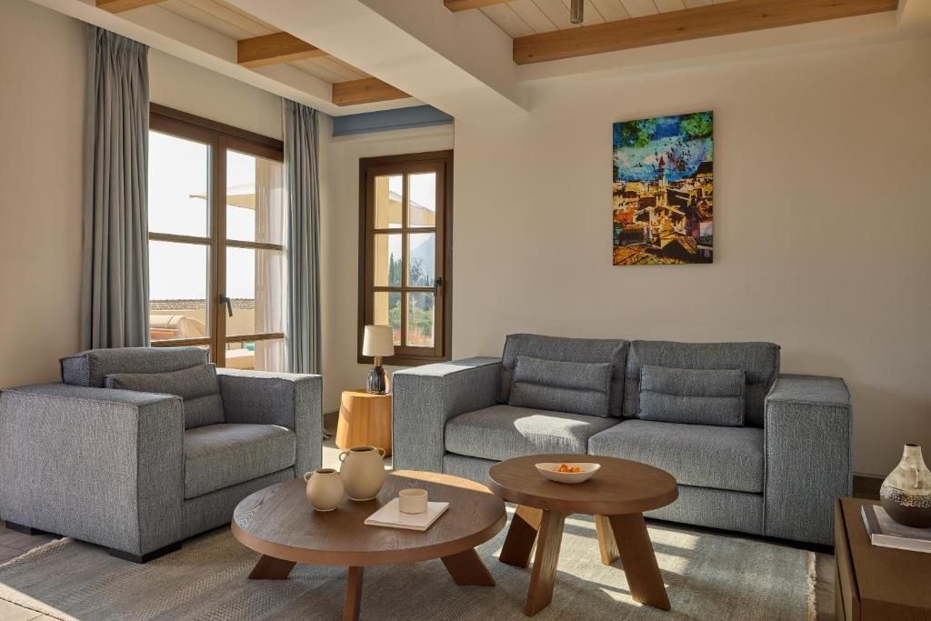 Sitzgruppe mit graublauer Couch, Sessel und zwei runden Holztischen vor den Fenstern.