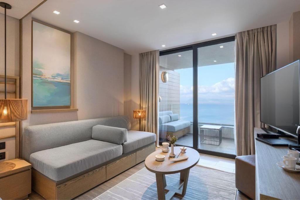Wohnbereich einer Suite mit grauem Sofa, Holztisch und Glasfront zum Balkon mit Meerblick.