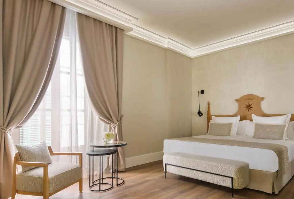 Hotelzimmer mit Doppelbett, Sitzbank, Sessel und zwei kleinen Beistelltischen am Fenster