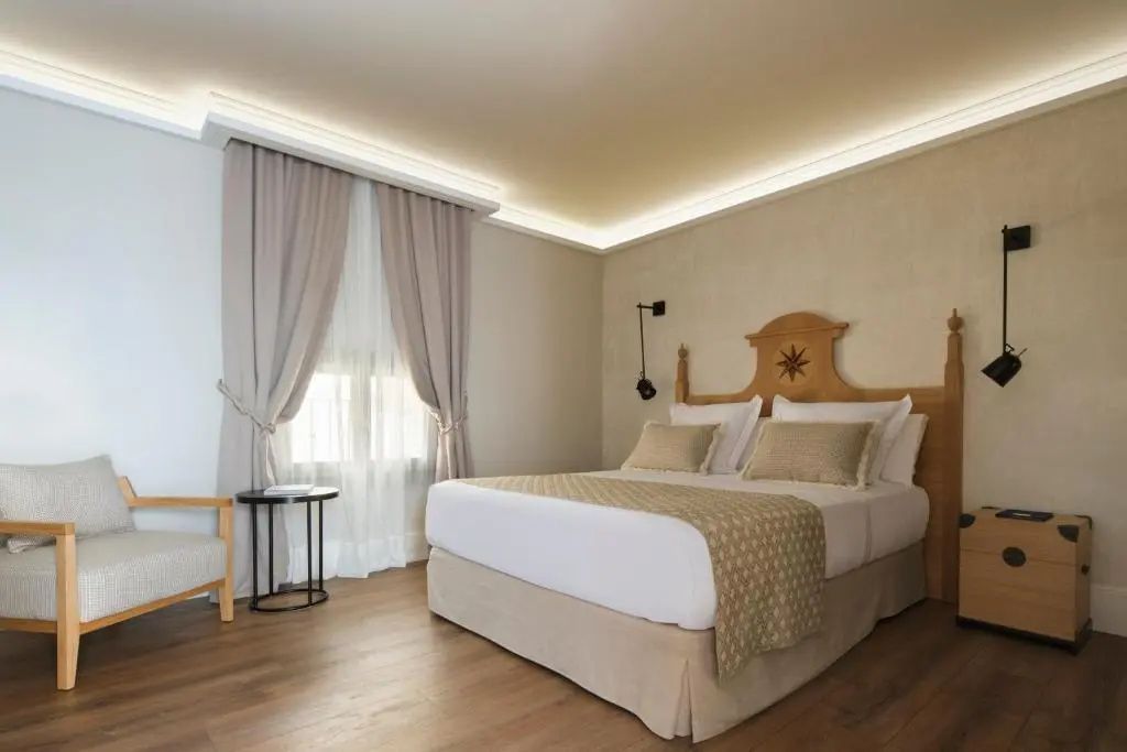 Doppelbett mit hellem Kopfteil aus Holz, beigefarbener Tagesdecke und Vorhängen im Hotelzimmer.