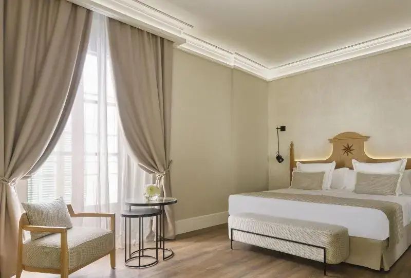 Hotelzimmer mit Sitzbereich am hohen Fenster, Vorhängen, Beistelltischen und Doppelbett.
