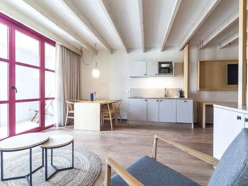 Offener Wohnbereich mit kleiner Küchenzeile, Esstisch, Holzbalkendecke und rotem Fensterrahmen.