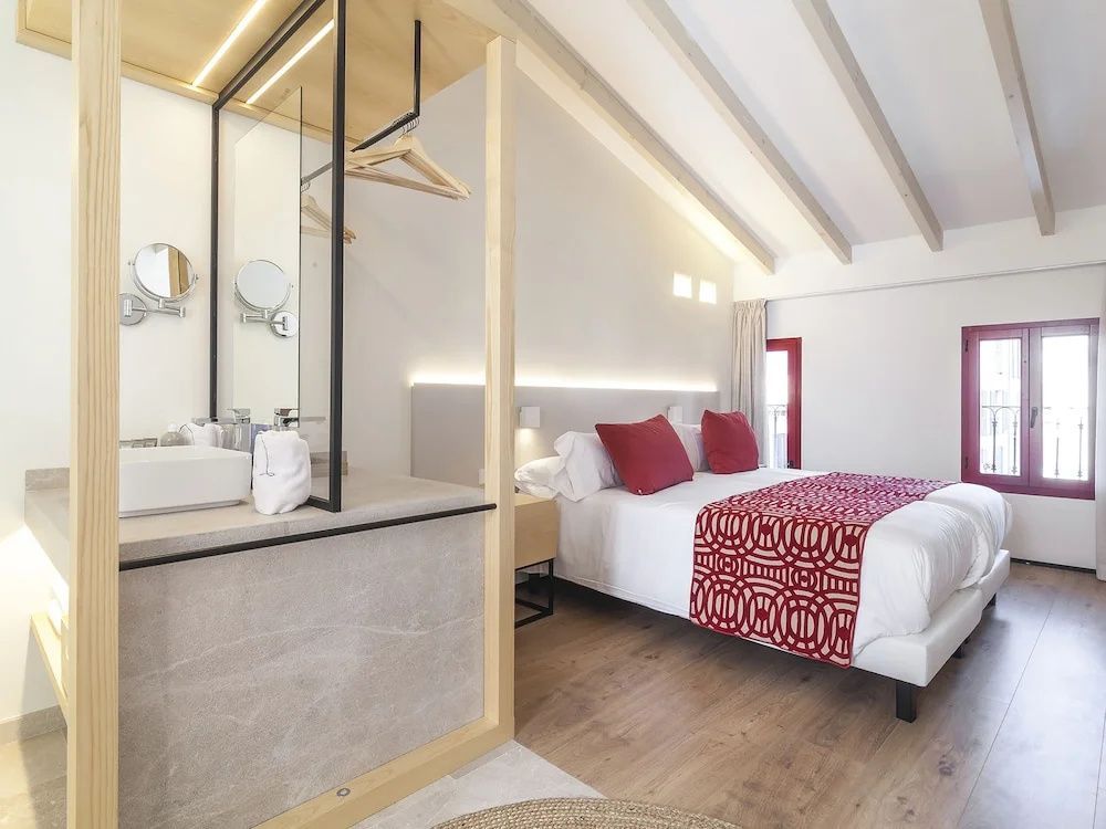 Helles Hotelzimmer mit sichtbaren Holzbalken an der Decke und rot dekoriertem Doppelbett.