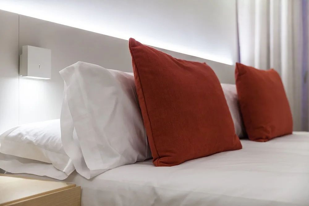 Nahaufnahme von weissen Kopfkissen und einem roten Dekokissen auf einem Bett mit Lichtleiste.
