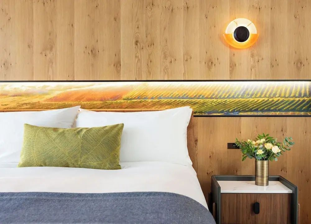Doppelbett mit weißer Bettwäsche, grünem Kissen und beleuchteter Rückwand aus Holz.