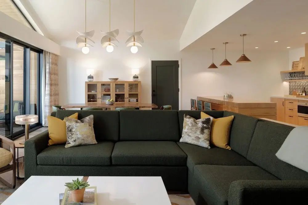 Dunkelgrünes Ecksofa mit gelben Kissen, weißer Couchtisch und Esstisch im Hintergrund.