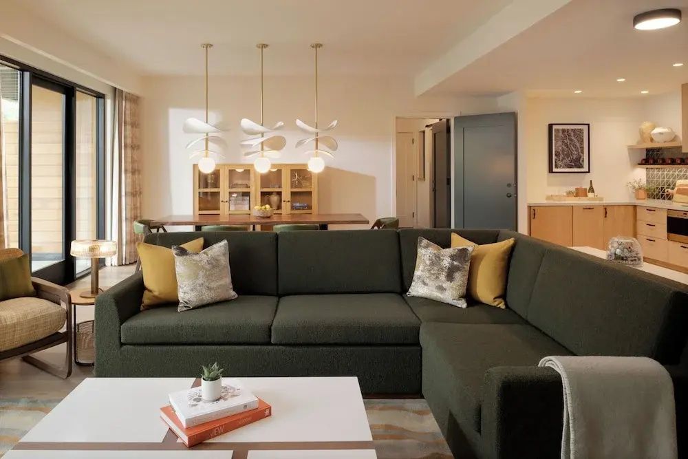 Modernes Ecksofa in Dunkelgrün mit Kissen, flacher Couchtisch und Holzmöbel im hellen Wohnbereich.