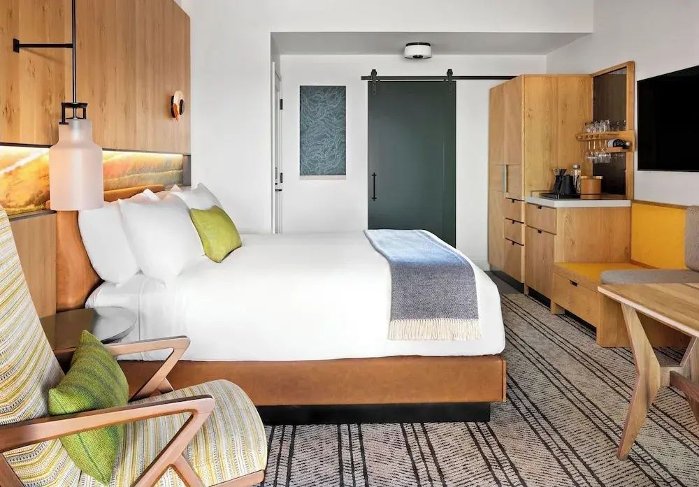 Hotelzimmer mit Doppelbett, Holzwand, Sessel, Schiebetür und gestreiftem Teppich.